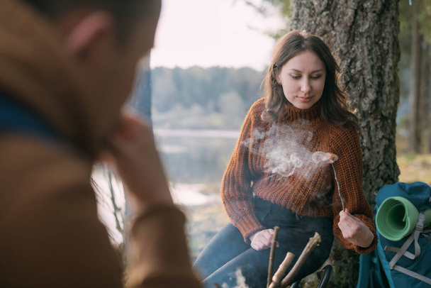 湖のほとりにある夫婦のフライドポテトマシュマロにテントがあります。森の中でたき火をしたハイキングキャンプ。男と女は熱い飲み物のマグカップで火による沈黙を楽しむ - 写真・画像