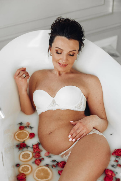 attractive pregnant woman relaxing in the bath with white water and a lot of oranges and flowers - Photo, Image