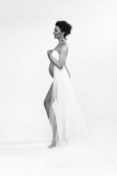 черно-белая фотография привлекательной беременной женщины, держащей белую ткань и стоящей в стороне от камеры - Фото, изображение