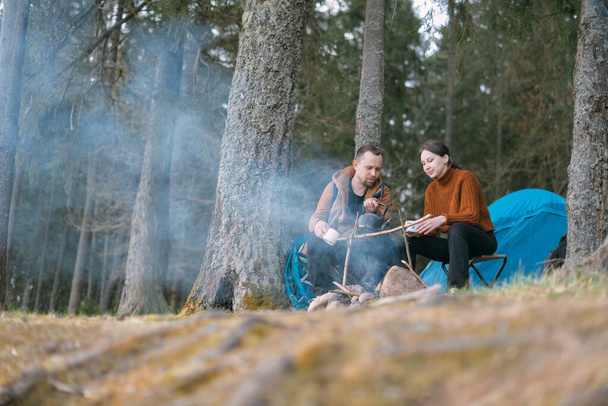 Una pareja de casados prepara comida en una fogata en la orilla del lago con una tienda de campaña. Campamento de senderismo con una hoguera en el bosque. Un hombre y una mujer disfrutan del silencio junto al fuego cerca de la tienda - Foto, imagen