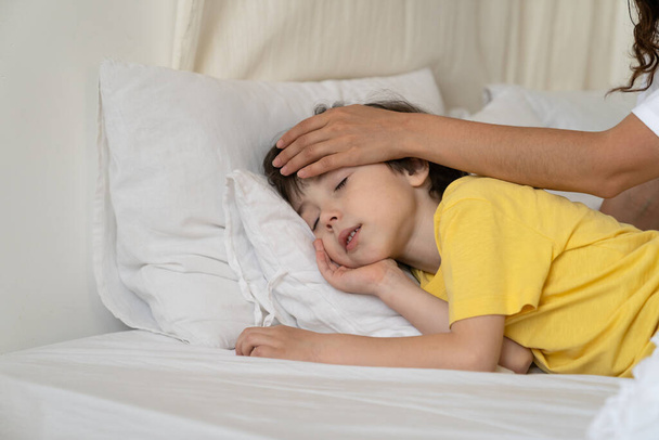 Huolehtiva äiti tarkistaa lapsen lämpötilan kosketa rauhallisen pojan otsaa nukkumassa sängyssä kuumeen ja influenssan jälkeen - Valokuva, kuva