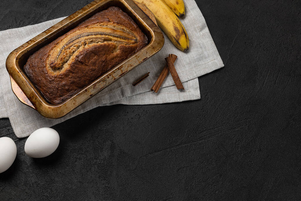 Ready fresh banana bread in rectangular baking dish with ingredients on dark background - Φωτογραφία, εικόνα