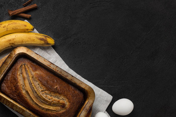 Ready fresh banana bread in rectangular baking dish with ingredients on dark background - Photo, Image