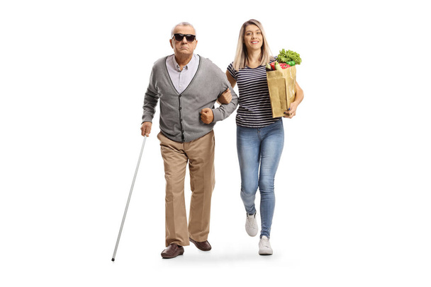 Full length portrait of a young woman carrying a grocery bag and helping an elderly blind man isolated on white background - Foto, Bild