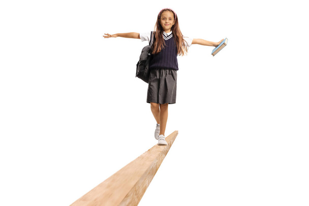 Schoolgirl in a uniform carrying a backpack and walking on a wooden beam isolated on white background - Fotografie, Obrázek