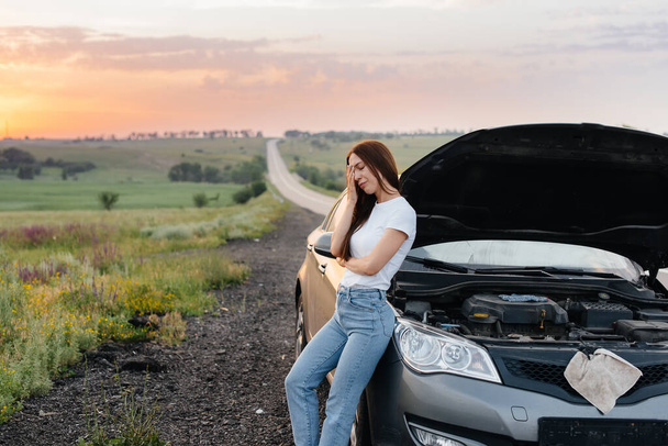 A frustrated young girl stands near a broken-down car in the middle of the highway during sunset. Breakdown and repair of the car. Waiting for help. - Photo, image