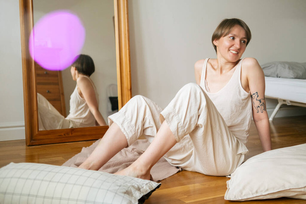 Lifestyle portrait of woman wearing linen pajama, sitting on floor and enjoying cozy home weekend. - 写真・画像