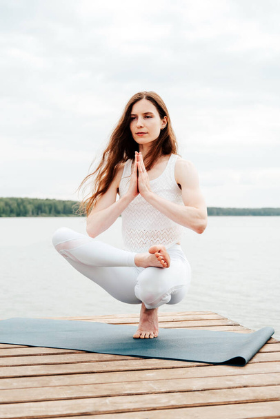 Beautiful young woman in white sports clothes is sitting on  wooden pier on lake in  balance yoga position with namaste gesture. - 写真・画像