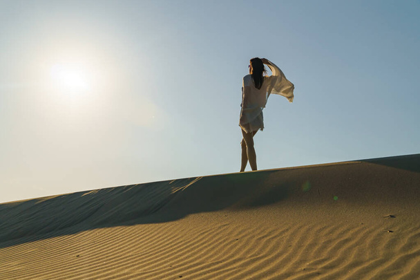 Millennial Woman standing on wavy sand dunes in desert landscape at golden sunset light and wind. - Fotó, kép