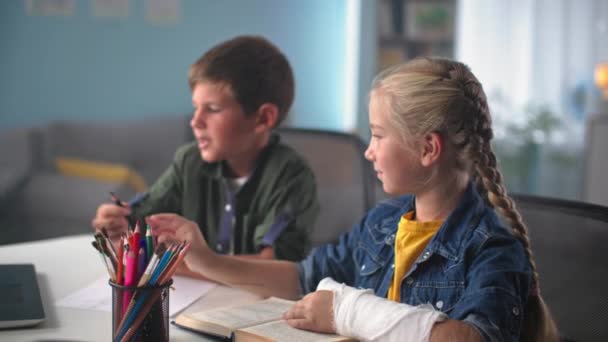casa escola, menina com gesso moldado na mão dá um lápis para o irmãozinho enquanto sentado à mesa durante uma lição de desenho - Filmagem, Vídeo