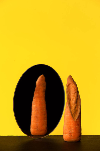 Zanahoria fea delante del espejo, reflejo, reverso. Concepto - reducción del desperdicio de alimentos orgánicos. Uso en la cocina de productos imperfectos. Hortalizas y frutas feas. - Foto, imagen