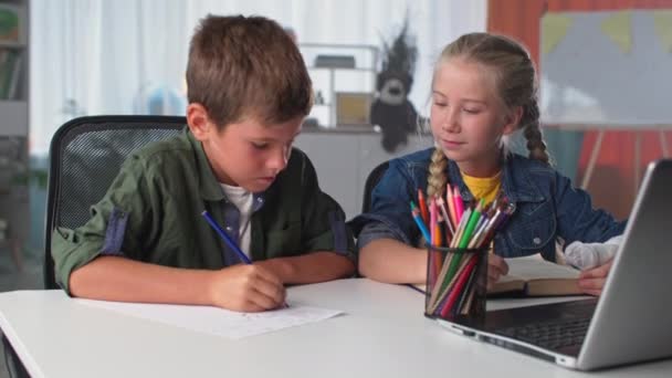 desenho lição em casa, menina bonito com um gesso moldado em sua mão dá lápis para o irmãozinho enquanto sentado na mesa com laptop no quarto - Filmagem, Vídeo
