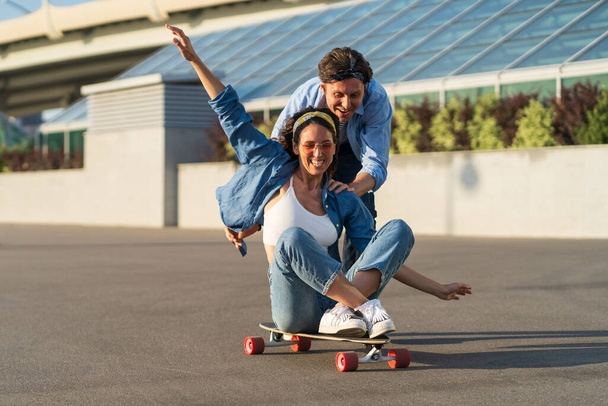 Couple have fun on longboard: male push back of joyful female sitting on skateboard and laughing - Foto, afbeelding