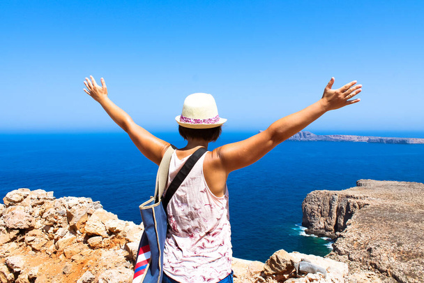 A tourist with a straw hat standing on a hill with her hands up to greet a blue sea at sunny summer day. Female traveler view from a back. Rest on a beach, the ocean. Travel the World. Explore Earth. - Foto, afbeelding