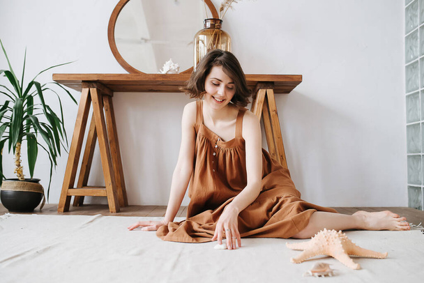 Curious sensual pretty woman sitting on the floor in a brown dress, touching a shell. Beautiful brunette with short hair indoors. - 写真・画像