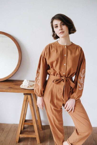Intriguing woman possing for a photo in a brown jumpsuit, looking at the camera. Beautiful brunette with short hair indoors, next to a table with a round mirror - Foto, afbeelding