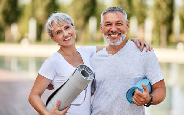 Lächelndes Seniorenpaar mit Gymnastikmatten im Park, positiver reifer Mann, der seine ältere Frau nach einem Fitness- oder Yoga-Kurs in der Natur umarmt. Wellness und gesunder Lebensstil im Ruhestand - Foto, Bild