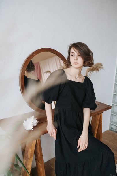 Pretty languid woman in a black dress leaning on a table with a round mirror. Beautiful brunette with short hair indoors. - Photo, Image