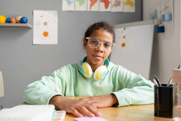 Urocza uczennica rasy mieszanej w okularach i odzieży casualowej siedząca przy biurku - Zdjęcie, obraz