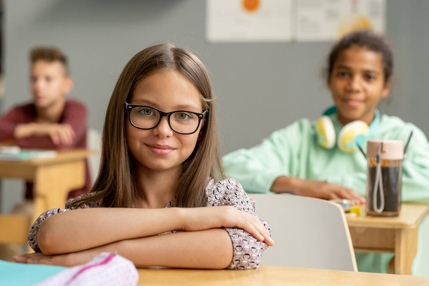 Roztomilá školačka v brýlích, dívá se na tebe za stolem - Fotografie, Obrázek