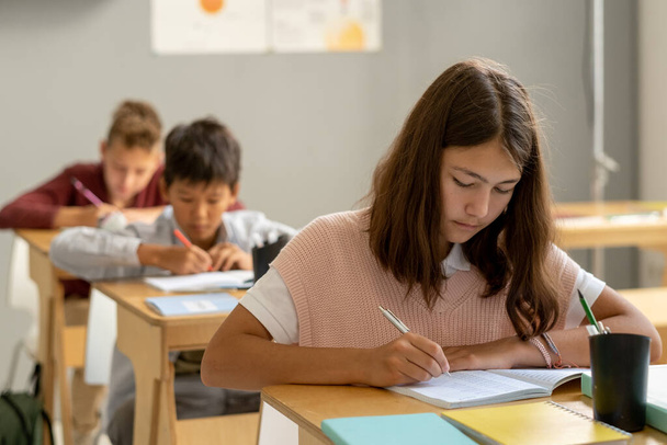 Aranyos iskoláslány jegyzetel a másolóban, miközben az asztal fölé hajol. - Fotó, kép