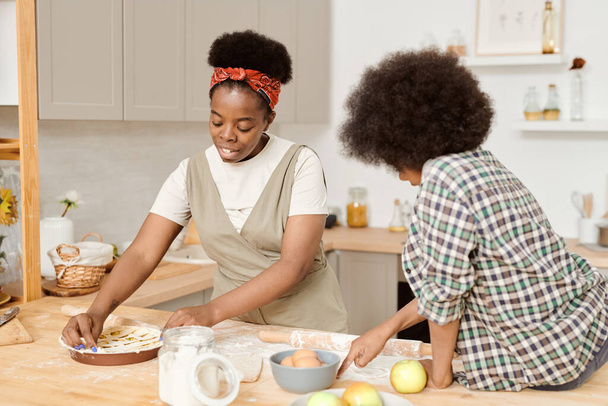 Young African female cooking apple pie with her cute son sitting near by - Photo, Image
