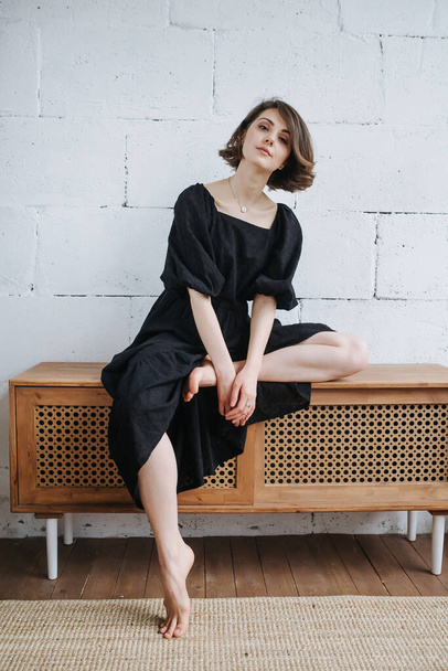Eccentric laguid woman sitting on a low cupboard in a modest black dress. With one leg up. Beautiful brunette with short hair indoors. - Фото, изображение