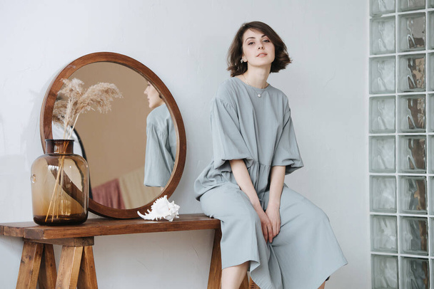 Portrait of a young woman in a grey dress sitting on a table with a round mirror. Her profile reflecting in the mirror. Beautiful brunette with short hair indoors. - Foto, immagini