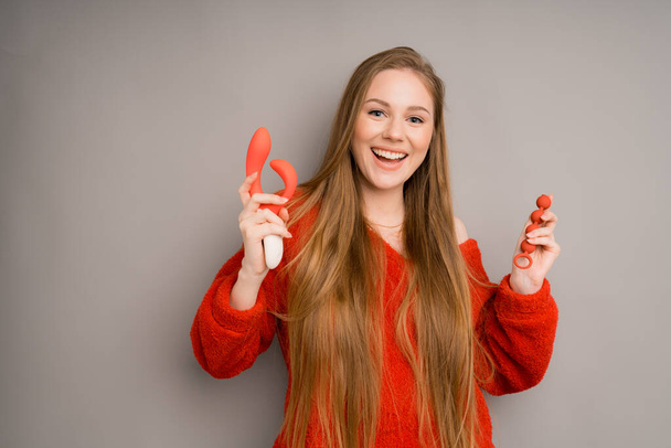 An attractive girl in a sweater depicts the emotion of happiness, smiling.holding a red dildo, vaginal balls in her hand - Zdjęcie, obraz