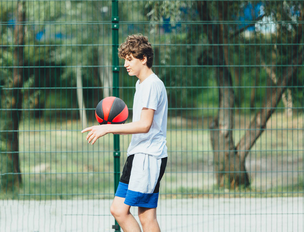 Милый подросток играет в баскетбол на городской площадке. Мальчик держит баскетбольный мяч в руках снаружи. Активная жизнь, хобби, спорт для детей - Фото, изображение