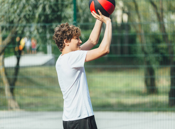 Милый подросток играет в баскетбол на городской площадке. Мальчик держит баскетбольный мяч в руках снаружи. Активная жизнь, хобби, спорт для детей - Фото, изображение