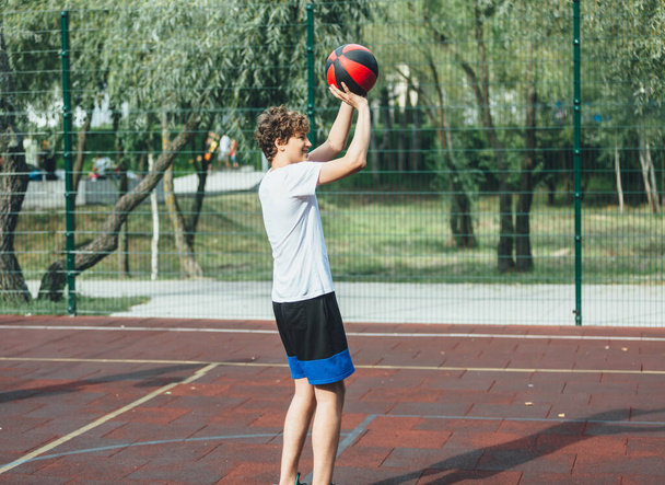 Leuke tiener speelt basketbal in een speeltuin. Een jongen houdt een basketbal in zijn handen buiten. Actief leven, hobby, sport voor kinderen - Foto, afbeelding