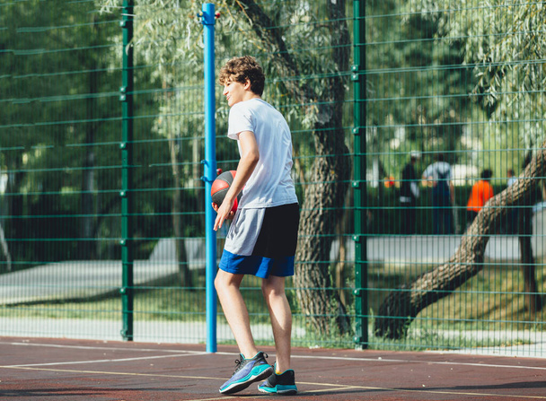 Aranyos tinédzser kosarazik egy városi játszótéren. Egy fiú egy kosárlabdát tart a kezében odakint. Aktív élet, hobbi, sport gyerekeknek - Fotó, kép