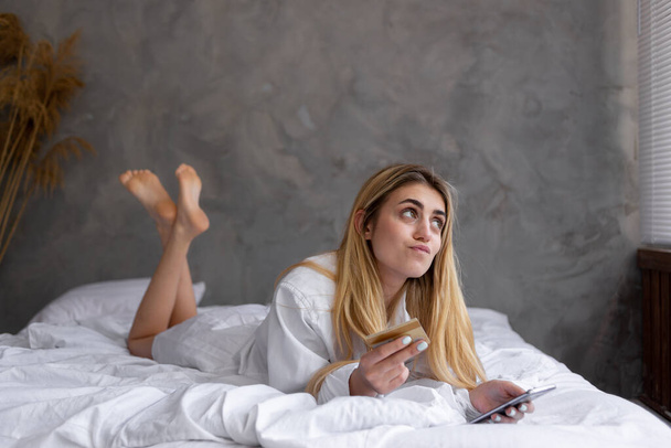 Retrato de una joven hermosa chica caucásica acostada en la cama en casa, de compras en línea. Utiliza una tarjeta de crédito y piensa en una elección difícil con su smartphone. Copiar espacio. - Foto, Imagen