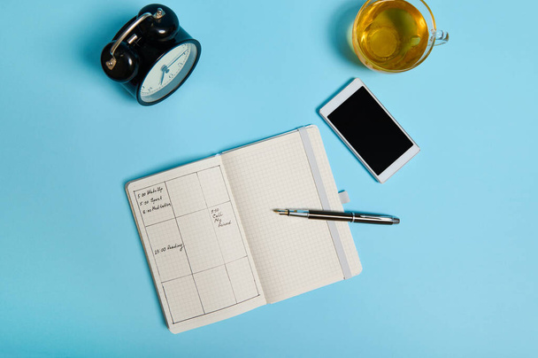 Flat lay of open agenda with schedule, smartphone, ink pen, glass cup of tea and black alarm clock lie on blue surface. Color background with copy space . Time management, deadline, schedule concept - Φωτογραφία, εικόνα