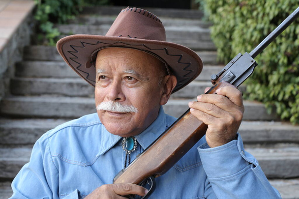 retrato de primer plano del hombre mayor guapo en camisa y sombrero de vaquero sosteniendo rifle al aire libre - Foto, Imagen