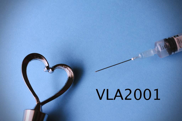 Primer plano de una aguja y un corazón en un fondo azul claro con el texto Vla2001, nueva vacuna contra el coronavirus - Foto, Imagen