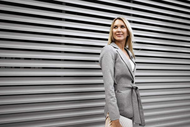 Portrait of young blonde businesswoman in gray suit walking in the city - 写真・画像