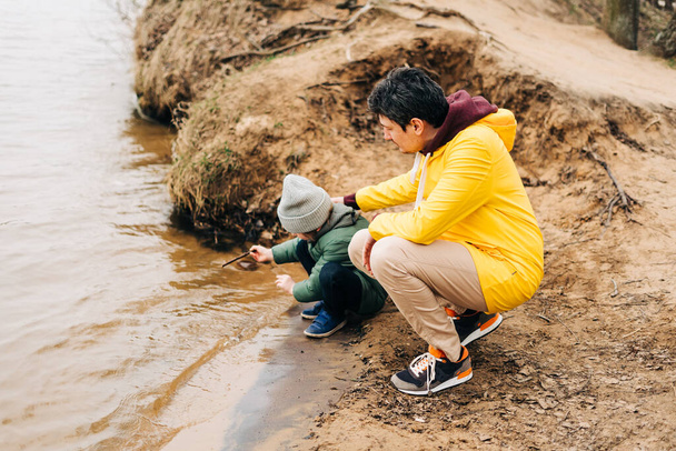Πατέρας και γιος στέκονται στην όχθη της λίμνης και παίζουν με το νερό. Ευτυχισμένη οικογένεια με παιδί παιδί αγόρι παίζει και να διασκεδάζουν σε εξωτερικούς χώρους κατά τη διάρκεια του φθινοπώρου πάρκο φόντο - Φωτογραφία, εικόνα