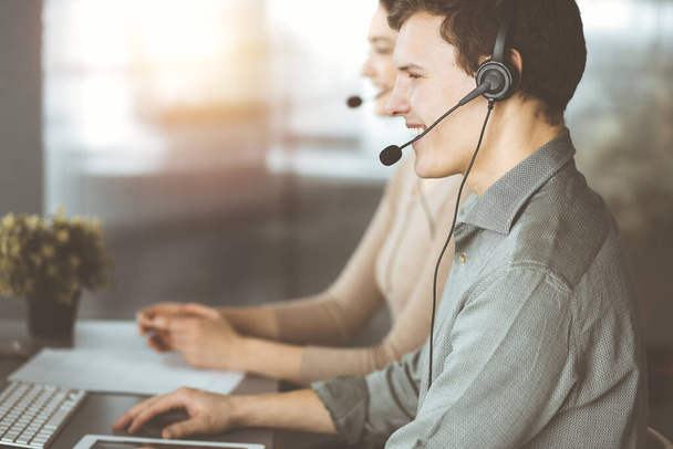 Young dark-haired guy with headsets is talking to a client, while sitting in sunny office. Call center operators at work - Foto, immagini