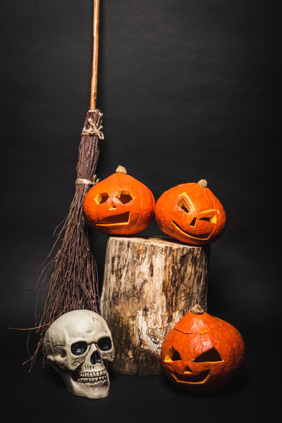 vassoura perto de crânio e laranja esculpidas abóboras no toco de madeira no preto - Foto, Imagem