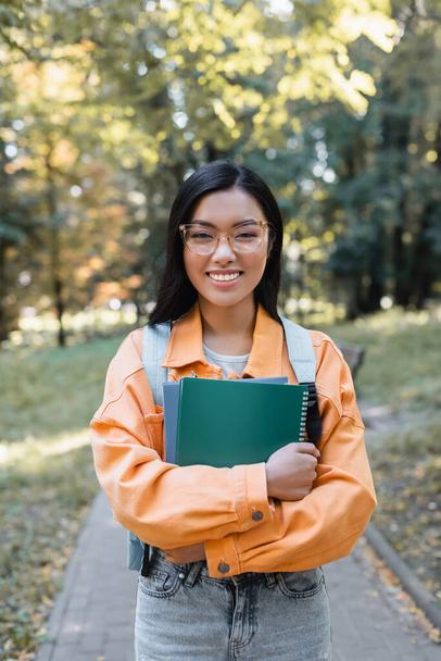 brunette asian student in eyeglasses smiling at camera while holding notebooks in park - Фото, зображення