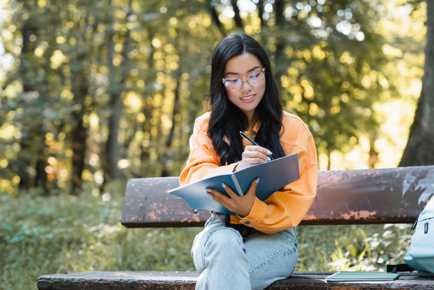 positieve aziatische student zitten op bank in park en schrijven in notebook  - Foto, afbeelding
