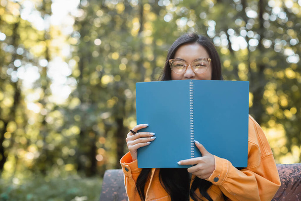 χαρούμενη ασιάτισσα μαθήτρια με γυαλιά ηλίου που σκιάζει το πρόσωπο με το βιβλίο αντιγράφων στο πάρκο - Φωτογραφία, εικόνα