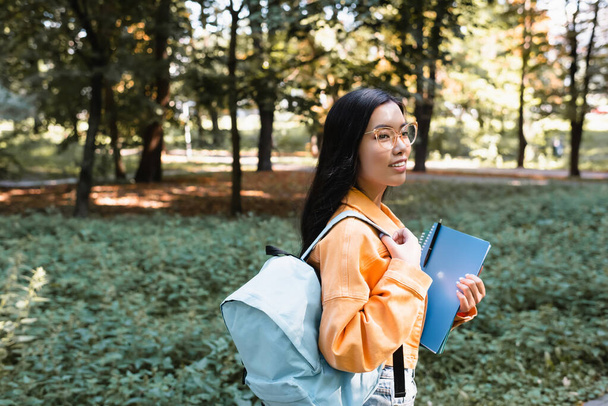 mladá asijská žena s batohem dívá pryč v parku, zatímco drží kopie knihy - Fotografie, Obrázek