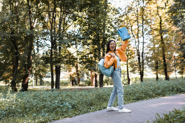 excited asian student waving notebooks while walking in park with backpack - Foto, Imagen