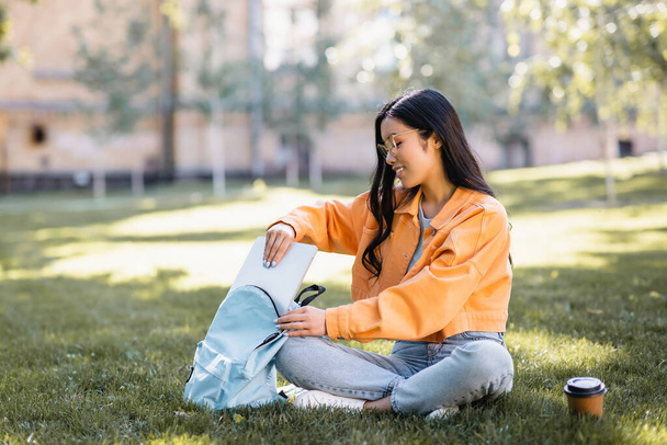 sonriente asiático mujer poner laptop en mochila mientras sentado en hierba con cruzado piernas - Foto, imagen