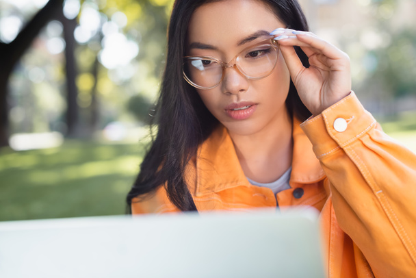 thoughtful asian woman touching eyeglasses while looking at blurred laptop outdoors - Zdjęcie, obraz