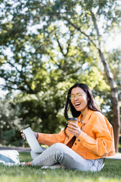 excited asian student with takeaway drink laughing while sitting on lawn with laptop - Photo, Image