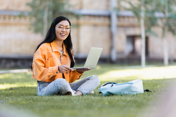 счастливая азиатка сидит на траве с ноутбуком и улыбается в камеру - Фото, изображение